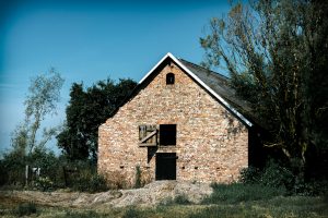 Brick barn - ProHort Ltd Abandoned brick barn with a steep triangular roof, small upper window, and surrounding trees against a blue sky.