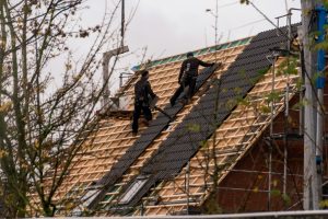 Roof replacement - ProHort Ltd Two workers walk on a steep, half-constructed roof with wooden battens and dark roof tiles, surrounded by scaffolding and equipment.
